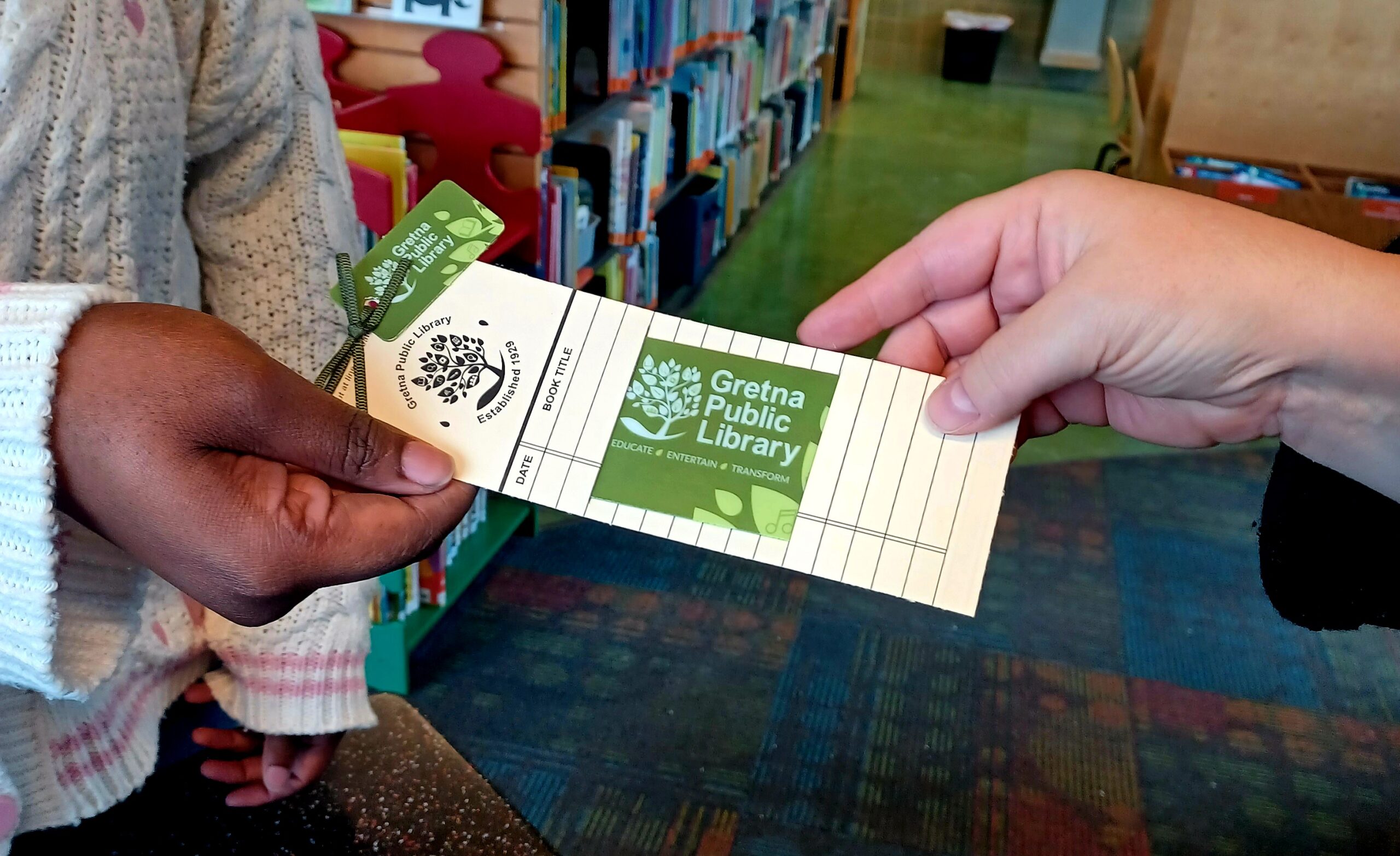 A closeup of two people's hands holding a new card bookmark. The card is passing between the two, presumable from a librarian to a patron.