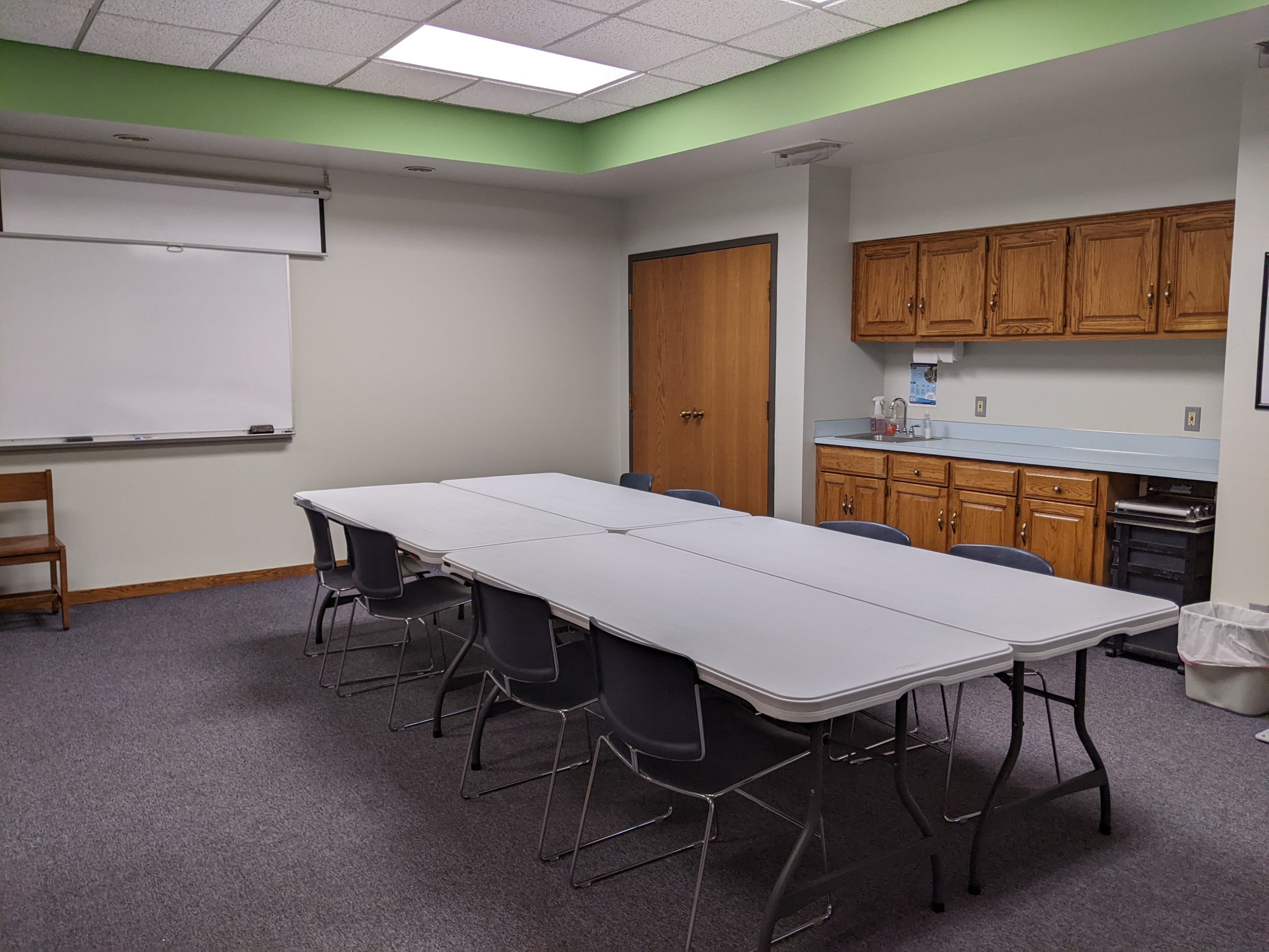 Image of the meeting room at Gretna Public Library. Features four folding tables, a wipe board, a sink, and chairs.