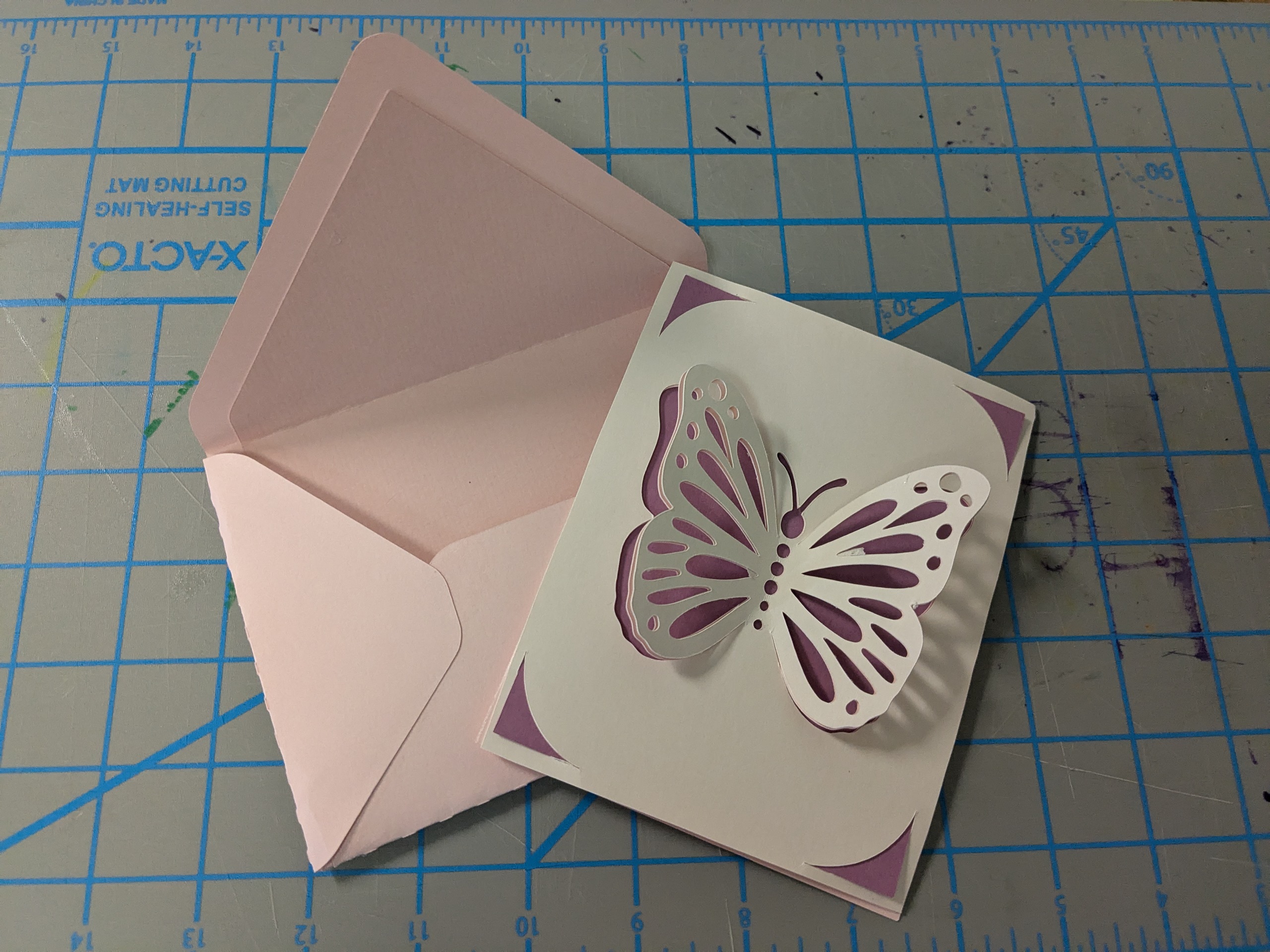 A card sits on a light pink envelope. The card is white, purple, and pink with butterfly wings popping out.